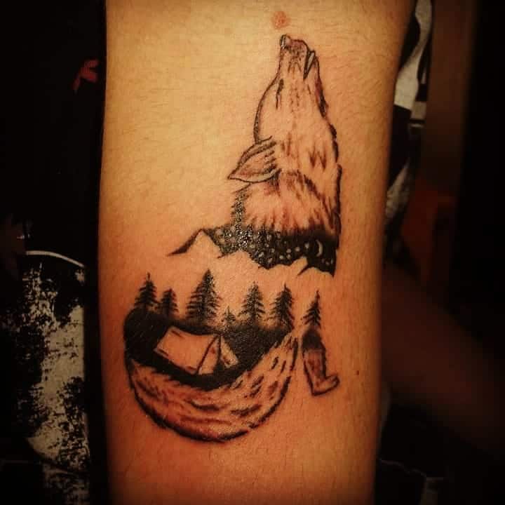 Tattoo of a howling wolf with trees, a tent, and mountains on an arm