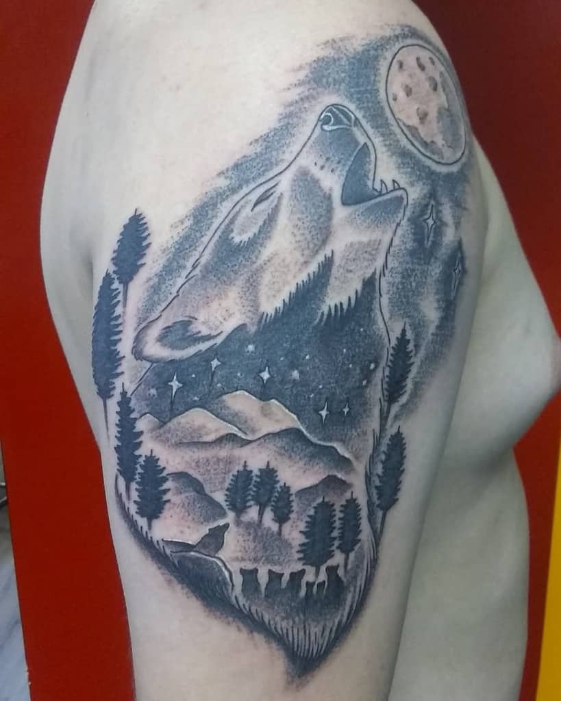 Tattoo of a howling wolf with trees, mountains, and a moon on a person's upper arm