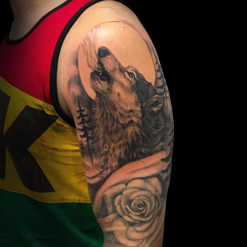 A striking tattoo of a lone wolf howling, surrounded by silhouettes of trees and a delicate rose, graces an arm