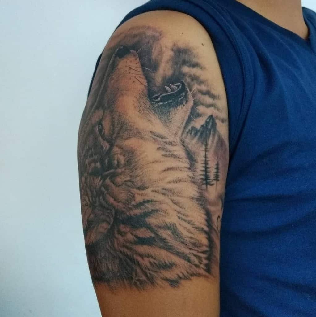 A stunning arm tattoo features a wolf howling amidst rugged mountains and towering trees