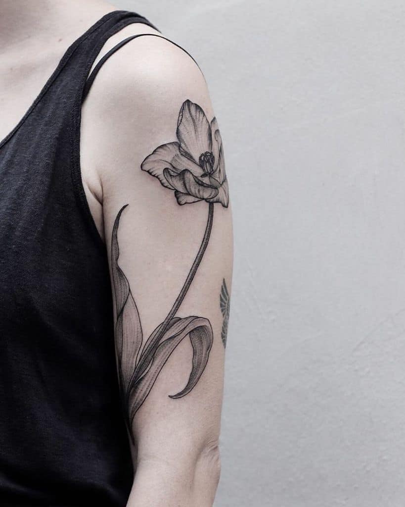 A detailed black-line tulip tattoo on the upper arm, featuring a large blooming flower with intricate shading and long, flowing leaves.