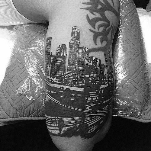 Detailed black ink tattoo of the Los Angeles skyline with freeway and palm trees on upper arm, featuring tribal design above.