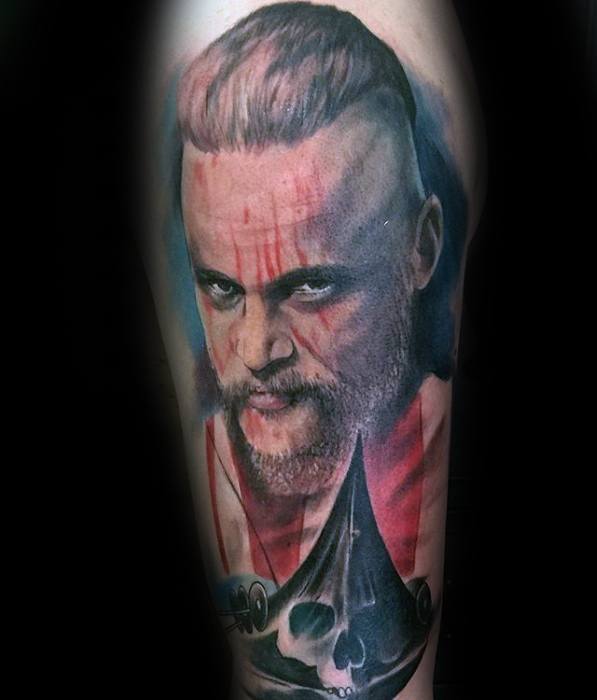 Realistic upper arm tattoo of Ragnar Lothbrok with blood streaks on his face and a skull below him.