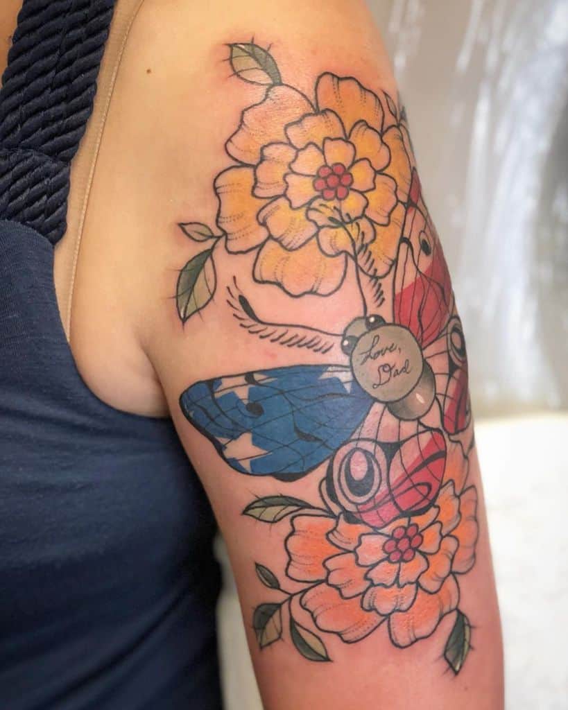Colorful tattoo featuring two orange marigolds, a red and blue moth with a locket inscribed 'Love, Dad,' and delicate green leaves