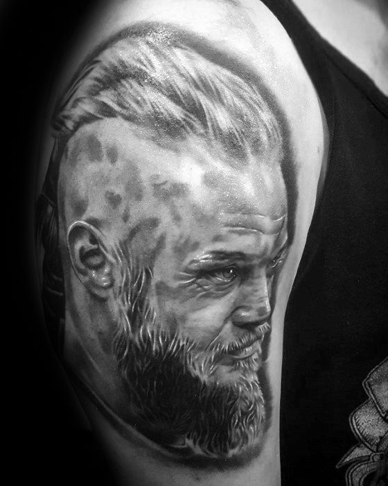 Realistic upper arm tattoo of Ragnar Lothbrok with detailed shading, braided hair, and intense gaze.