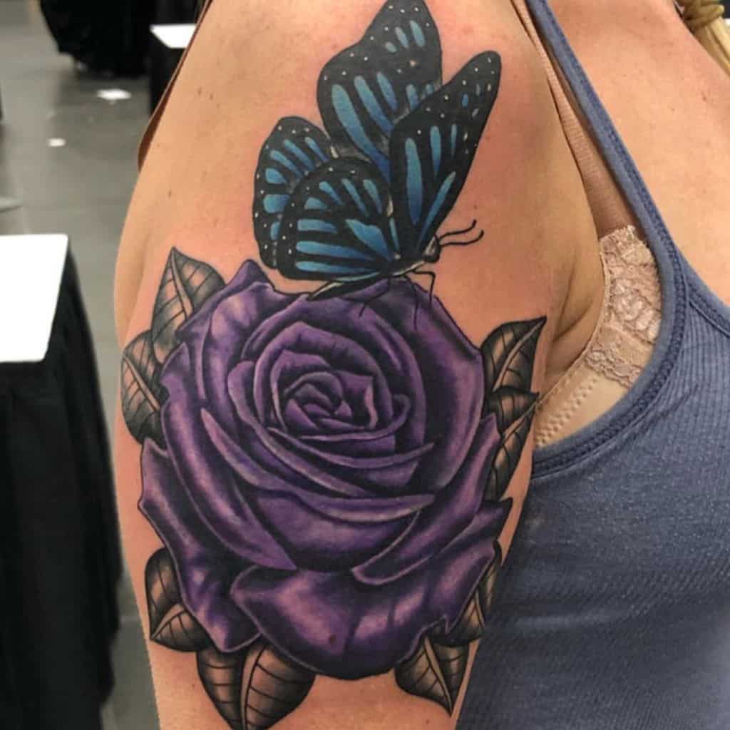 A stunning tattoo of a blue butterfly gracefully perched on a vibrant purple rose, artfully etched on the woman's upper arm