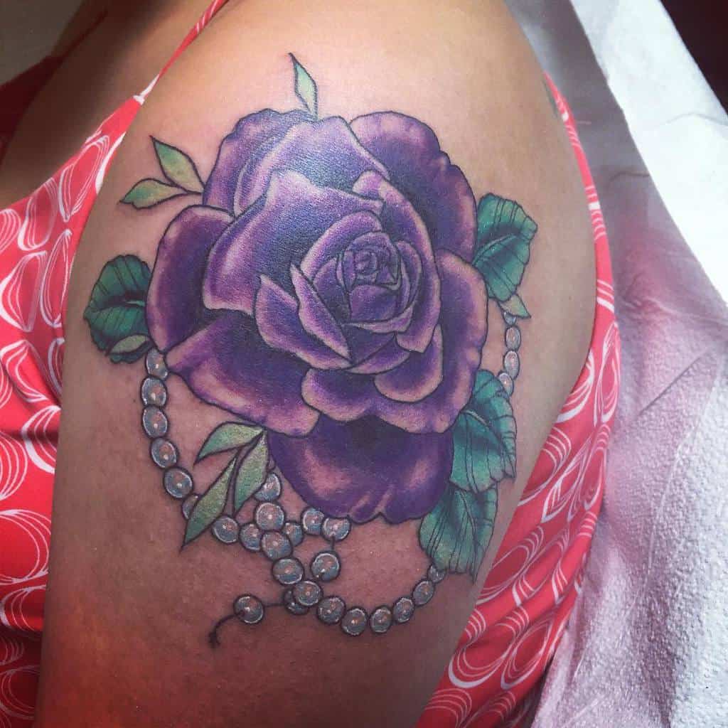 A stunning tattoo features a vibrant purple rose entwined with lush green leaves and a delicate string of pearls gracefully draped along the arm