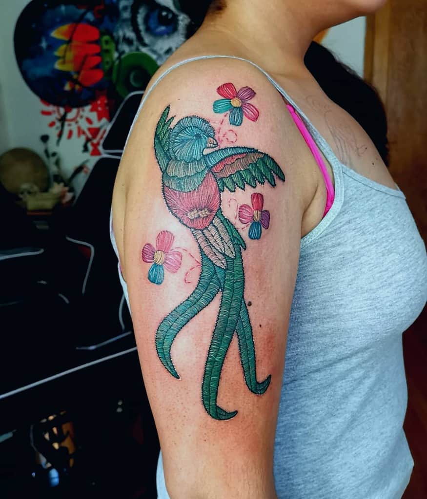 Colorful quetzal tattoo on the upper arm, featuring a stylized bird with intricate thread-like details, surrounded by vibrant flowers in shades of pink and blue
