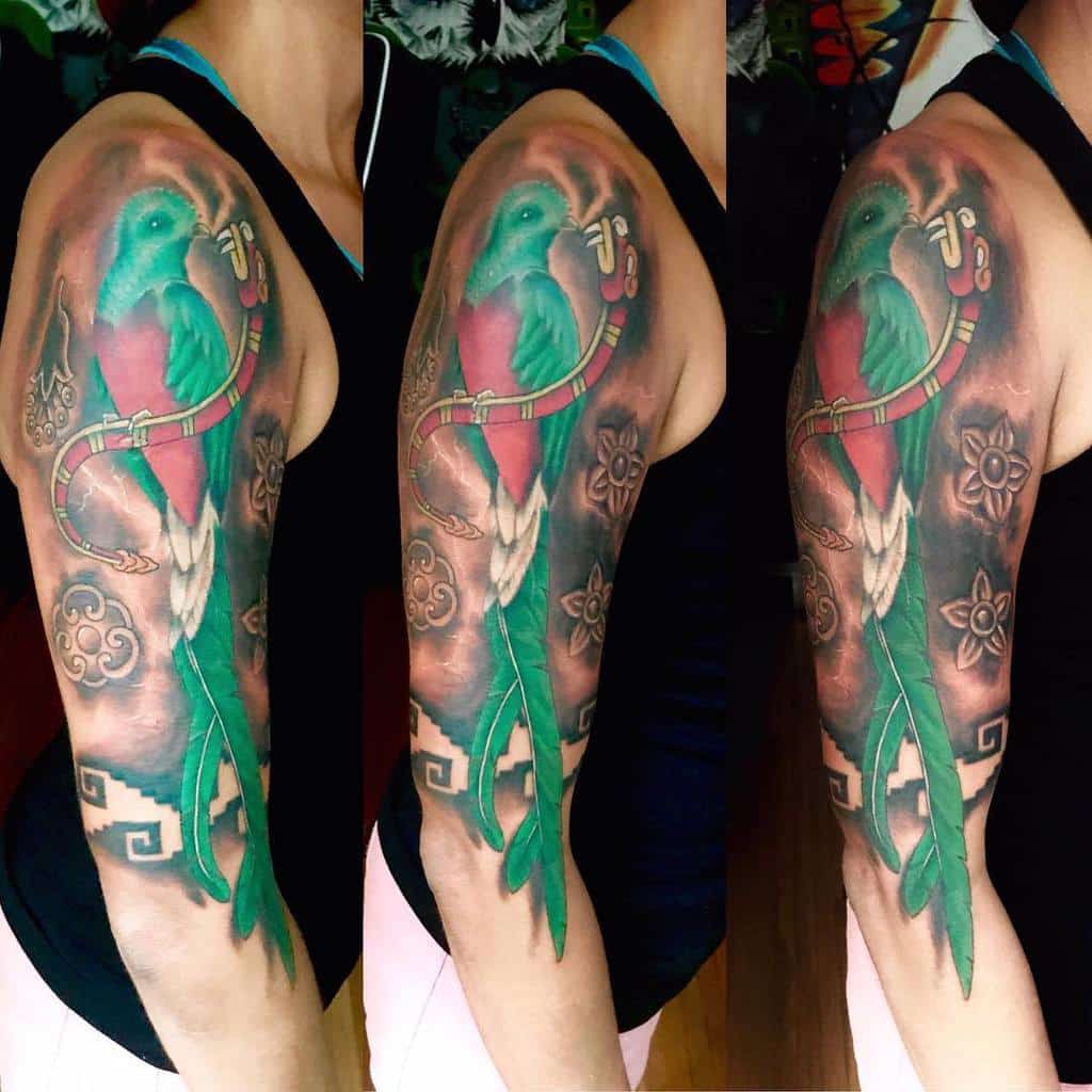 Full-sleeve quetzal tattoo on upper arm, featuring a vibrant red and green bird with long tail feathers, surrounded by Mayan-inspired designs