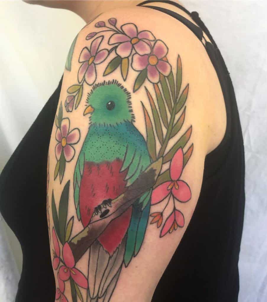 A vibrant quetzal bird tattoo on an arm, surrounded by pink flowers and green leaves