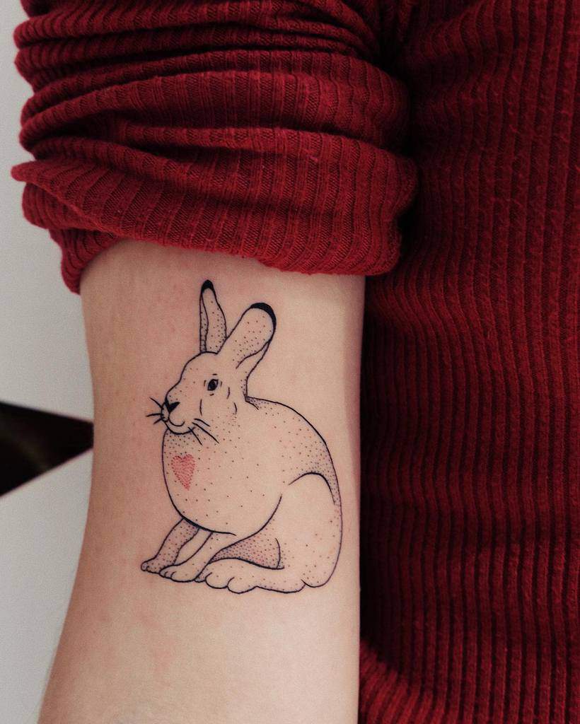 Simple line tattoo of a seated rabbit with a small heart on its chest, created using fine lines and dotted shading