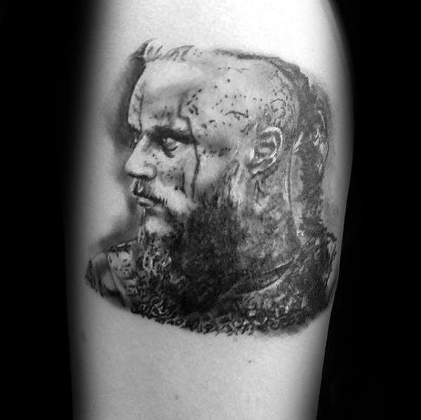 Ragnar Lothbrok portrait tattoo on upper arm, featuring battle scars, braided hair, and a fierce expression.