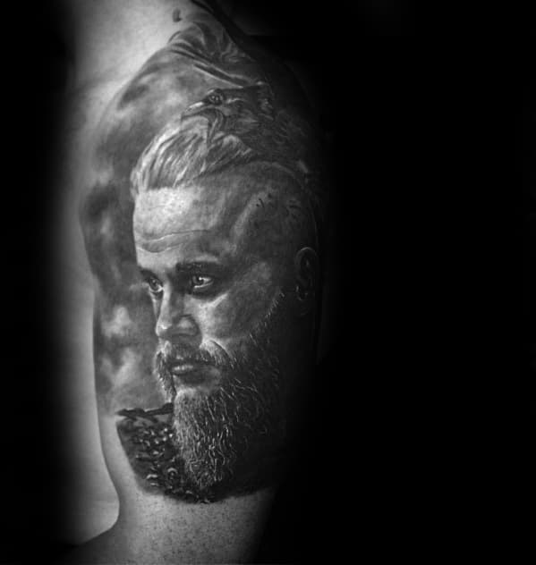 Black and gray Ragnar Lothbrok tattoo on upper arm, featuring his profile with detailed beard