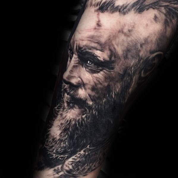 Detailed Ragnar Lothbrok tattoo on the forearm, depicting his profile with a long beard and scars.