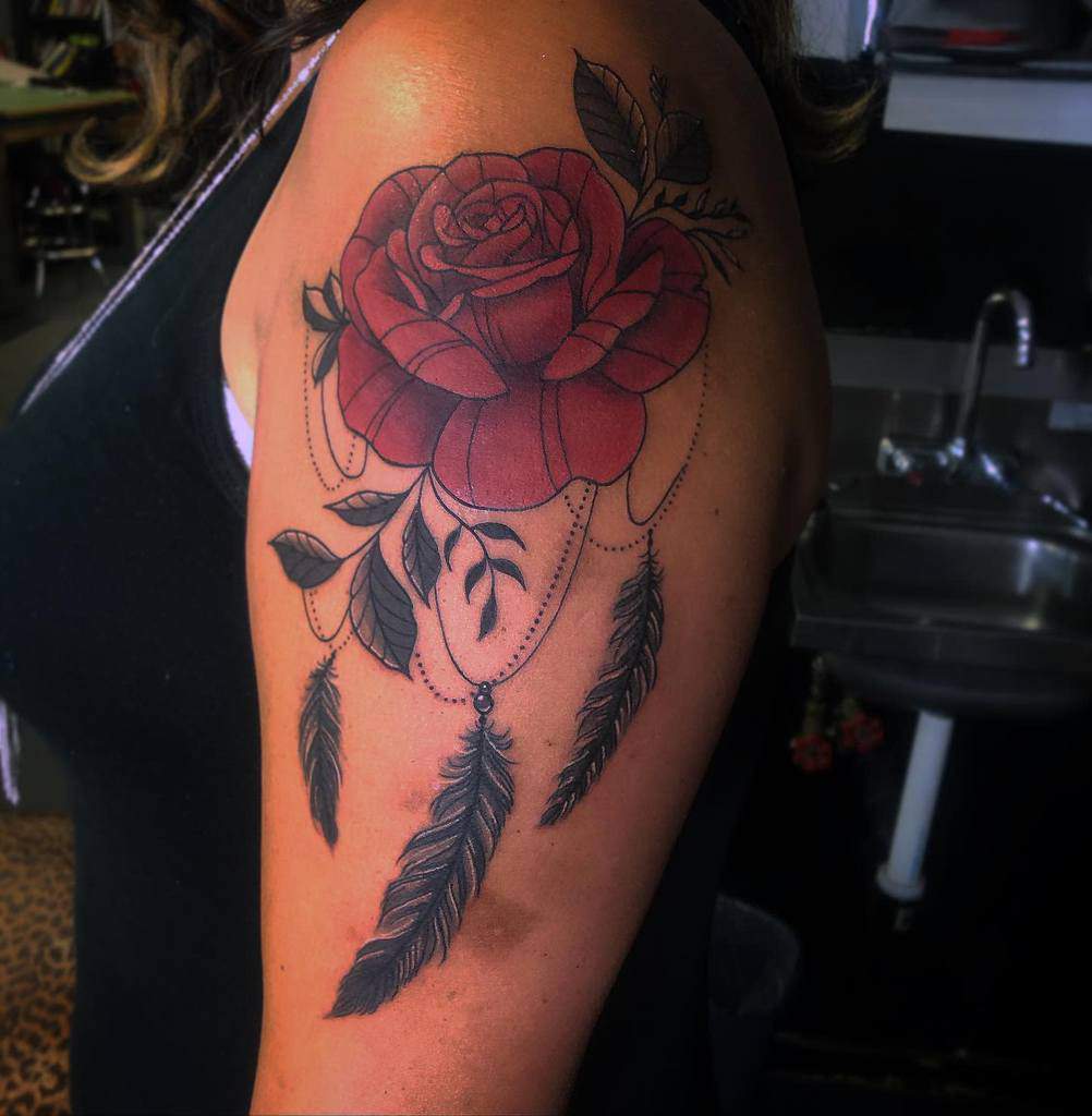 A striking tattoo of a red rose elegantly adorns a person's upper arm, complemented by delicate feathers that gracefully dangle beneath it