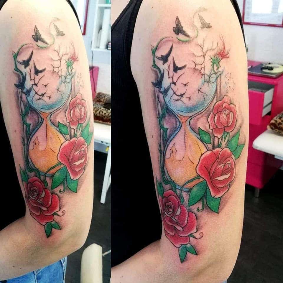 Vibrant tattoo featuring an hourglass surrounded by red roses and green vines, with flying birds symbolizing time and freedom