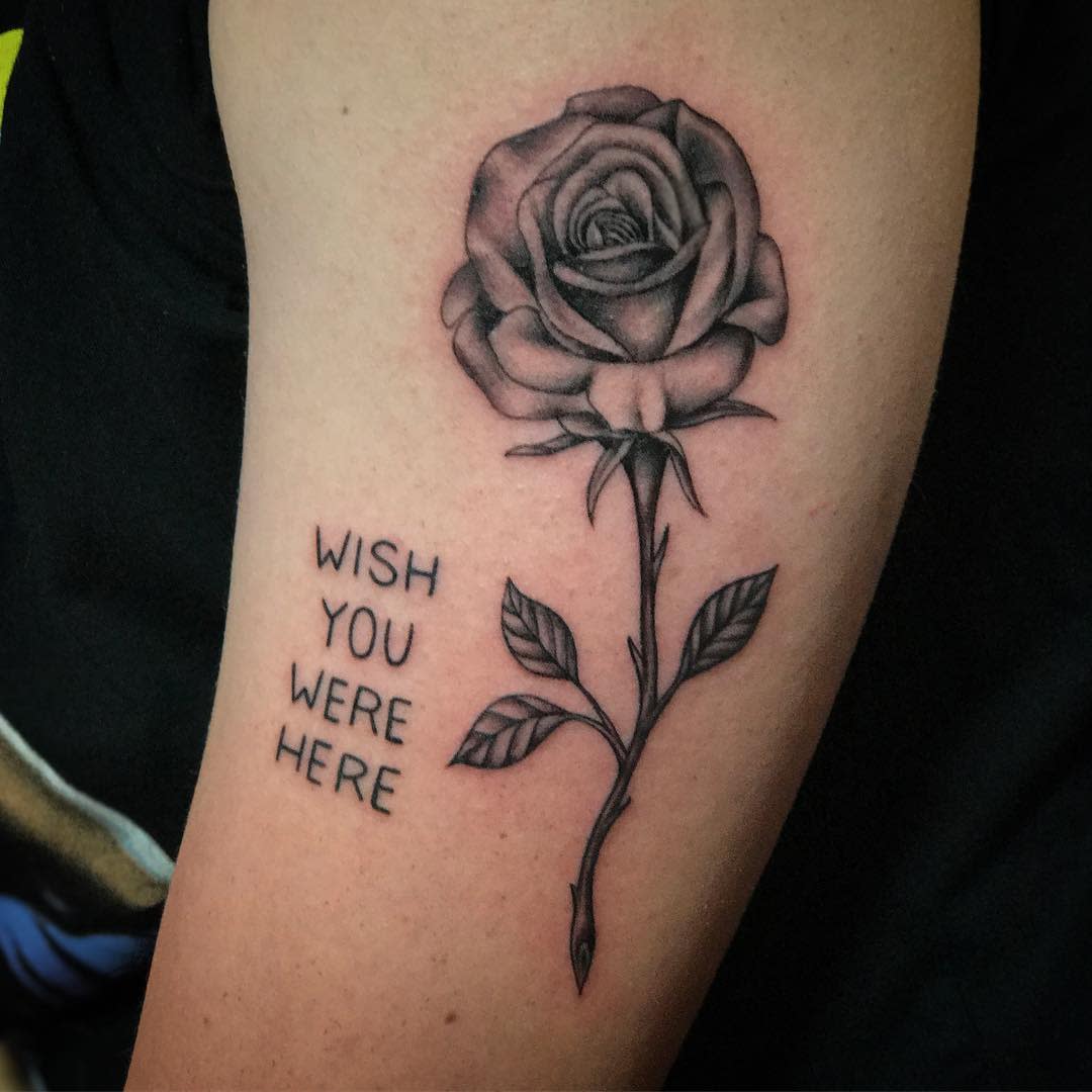 A delicate rose tattoo, complete with a graceful stem, wraps around a person's arm, bearing the heartfelt words "Wish you were here"