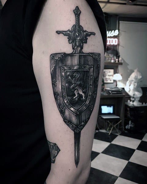 Black and grey tattoo of a sword with a detailed shield at the center, featuring a lion emblem, placed vertically on the upper arm.