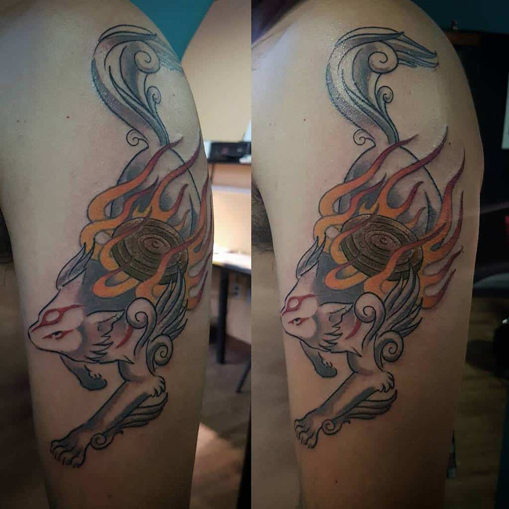 Dynamic Okami tattoo of Amaterasu on the arm and shoulder, featuring flowing divine flames and intricate swirling details in bold colors