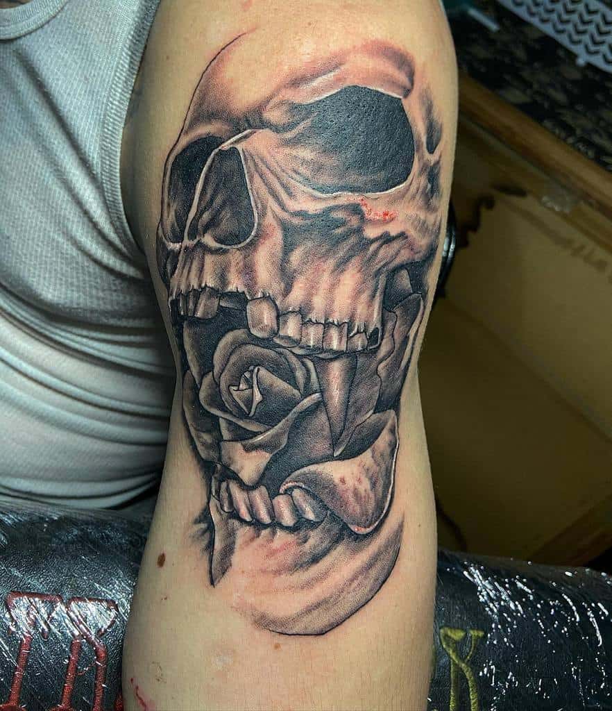 Realistic black and grey tattoo of a skull with a rose held between its teeth, featuring bold shading and intricate details on the arm.