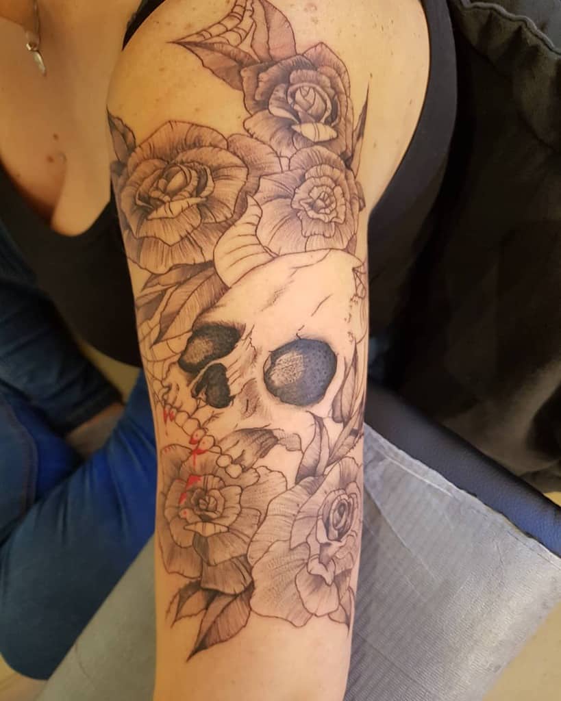 Black and grey tattoo of a skull surrounded by detailed roses and thorns, covering the upper arm and shoulder.