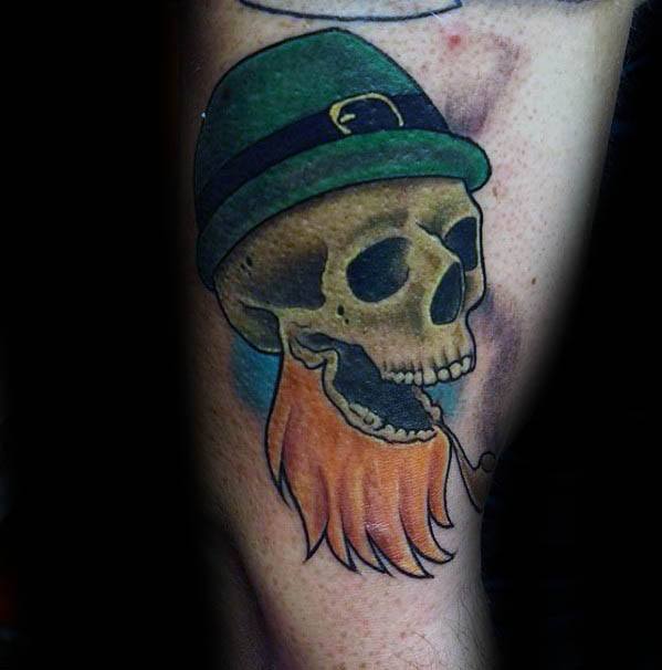 Tattoo of a leprechaun skull with a green hat, fiery orange beard, and a curved pipe hanging from its mouth, set against a soft blue background.