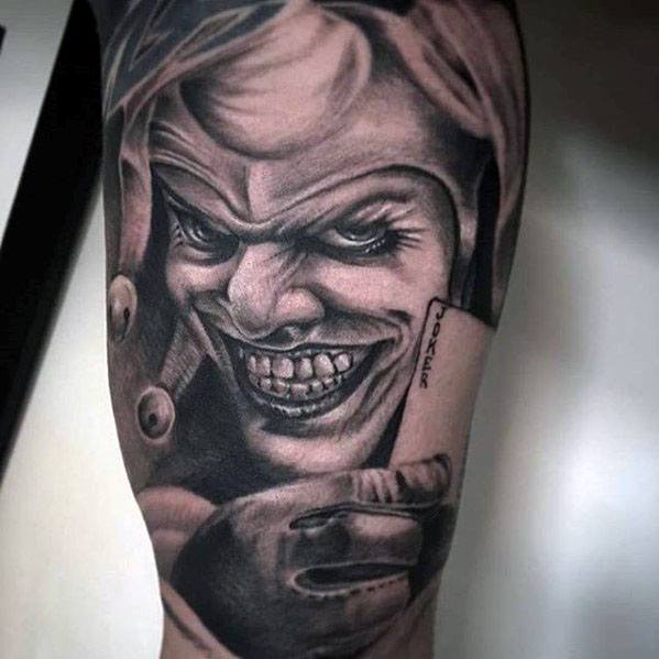 A detailed black and gray tattoo of a grinning jester holding a playing card with "Joker" written on it, showcasing intense facial features.