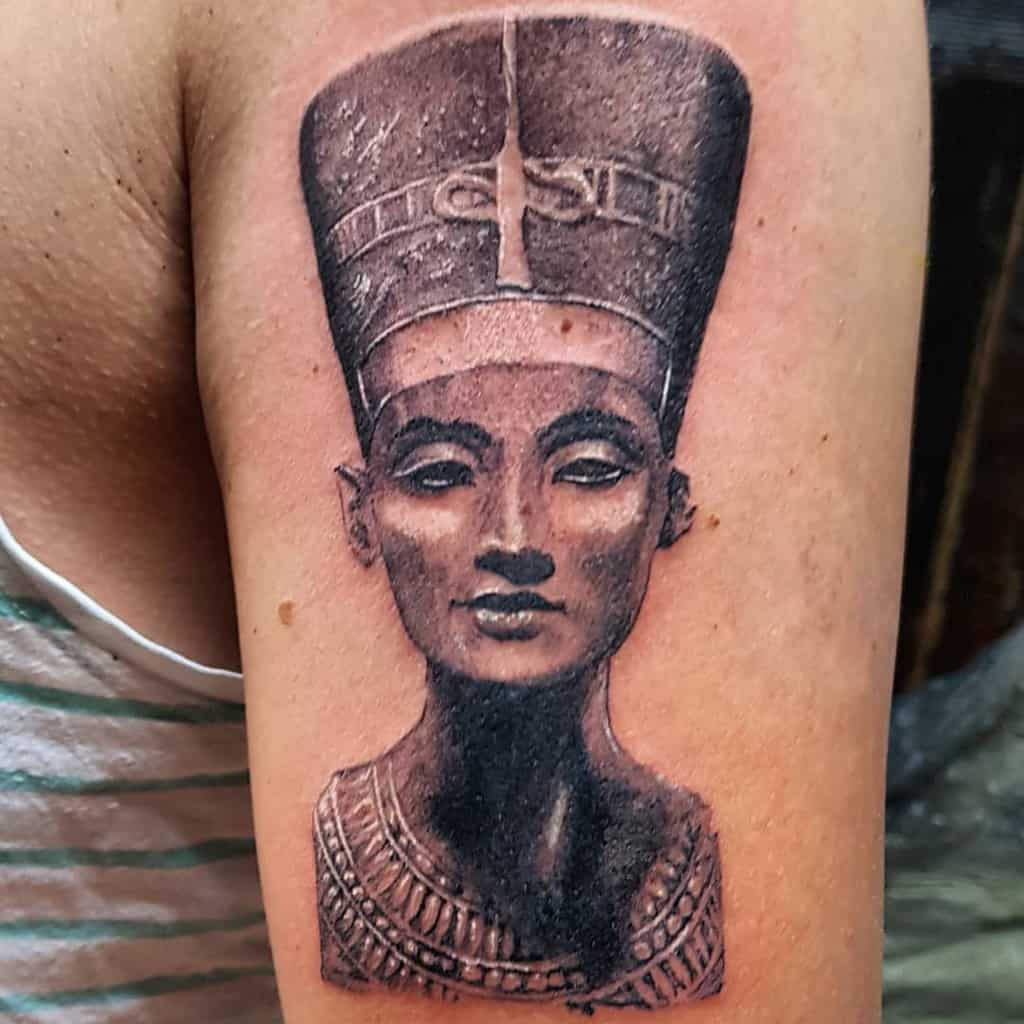 Tattoo of Nefertiti's bust on an upper arm, showcasing detailed facial features and elaborate headdress