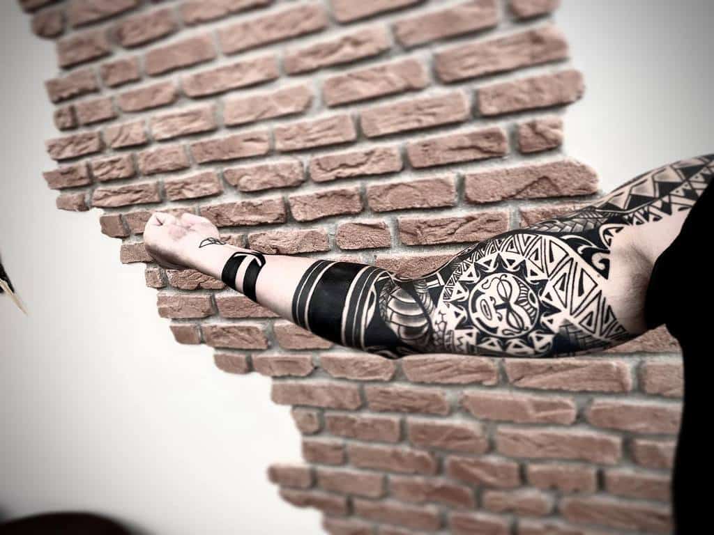 A bold black tribal tattoo covering the arm, featuring Tiki masks and geometric patterns