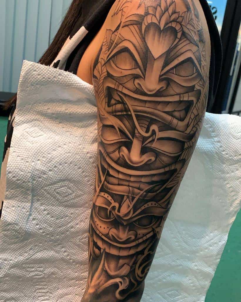 A detailed black-and-gray tattoo of multiple Tiki masks, with intricate shading and design, featuring fierce expressions and tribal patterns