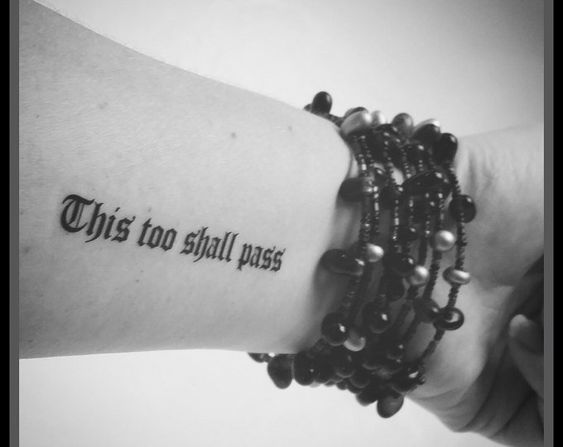 A "This Too Shall Pass" tattoo in bold font on the inside of a person's forearm, with beaded bracelets visible.