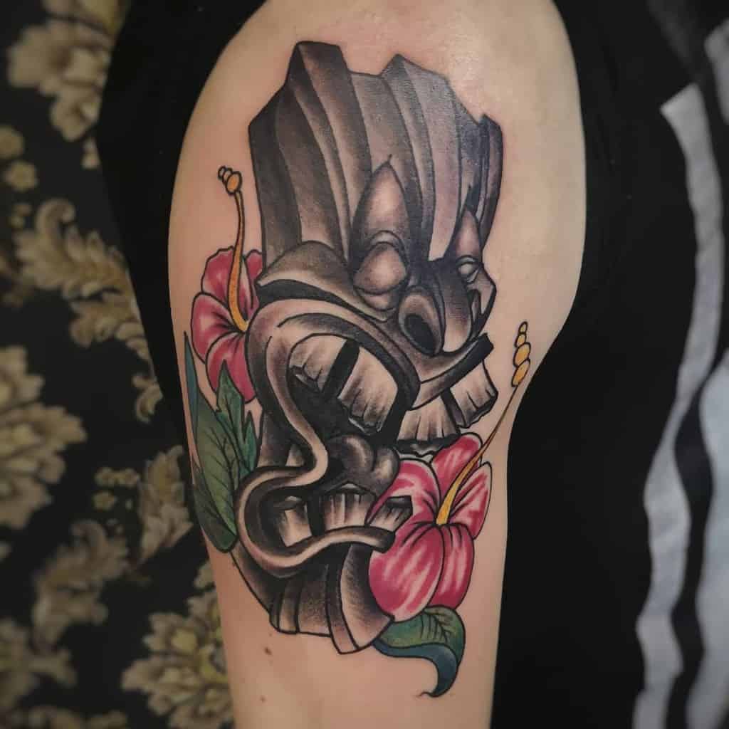 Bold black and grey Tiki tattoo featuring a carved Tiki mask, surrounded by vibrant pink hibiscus flowers and green leaves