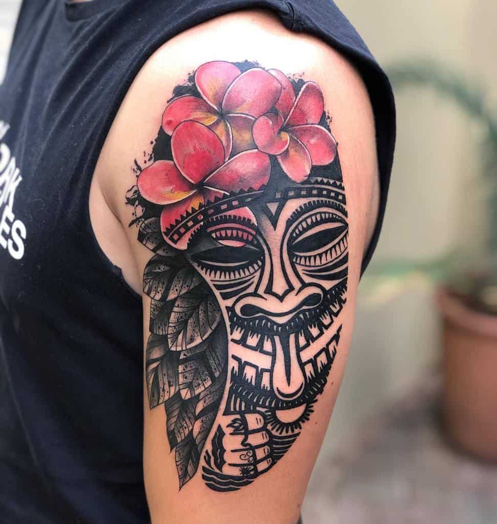 Vibrant Tiki tattoo on the shoulder featuring a detailed Tiki mask, adorned with red plumeria flowers, has blank ink leaves tattoo