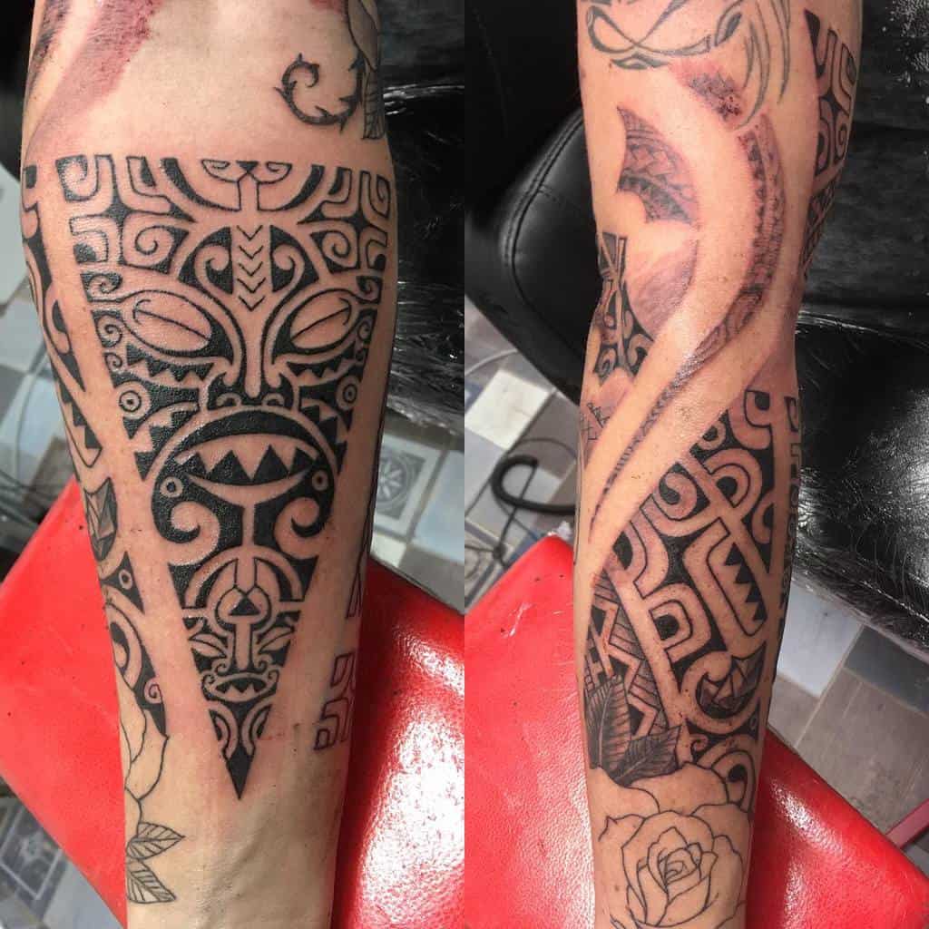 Intricate black and grey tribal tattoo featuring a bold Tiki mask surrounded by detailed geometric patterns and a spiral design, with a rose at the base