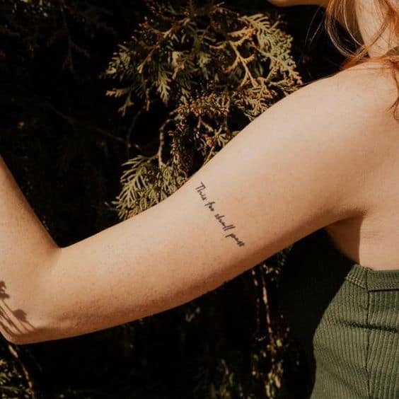 This image shows a tattoo on the upper arm of a person, reading "This too shall pass." The tattoo is in a simple and elegant script font, and the person is standing outdoors surrounded by greenery.