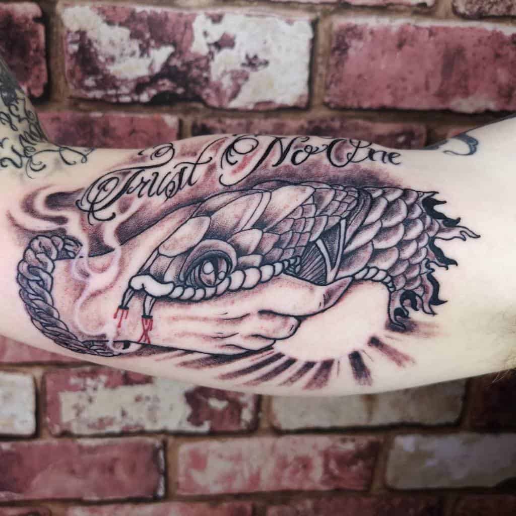 Detailed black and gray arm tattoo featuring a handshake where one hand is a snake, with the phrase 'Trust No One' above