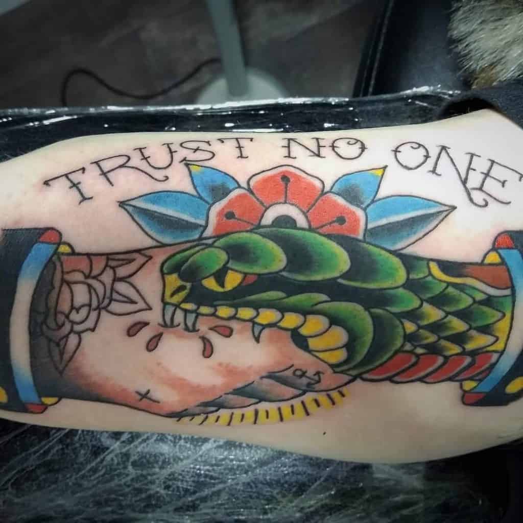 Vibrant arm tattoo of a snake biting a hand with blood drops, framed by a flower and the phrase 'Trust No One' above