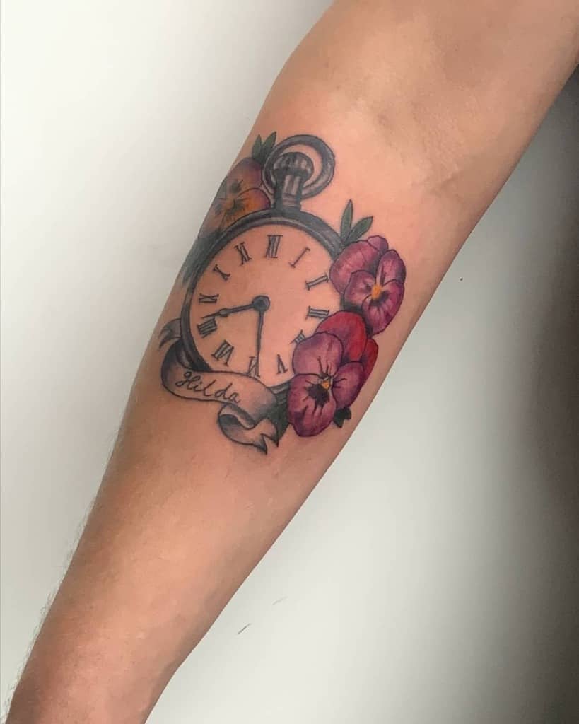 Vintage pocket watch tattoo with Roman numerals, surrounded by vibrant violet and red pansy flowers, and a ribbon with the name 'Hilda' for a personal touch