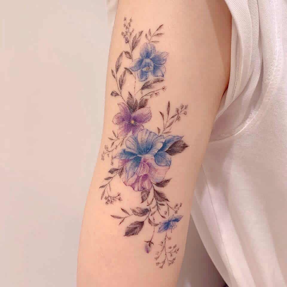 Delicate floral tattoo featuring purple and blue blossoms with fine leaves and stems, elegantly placed on the arm