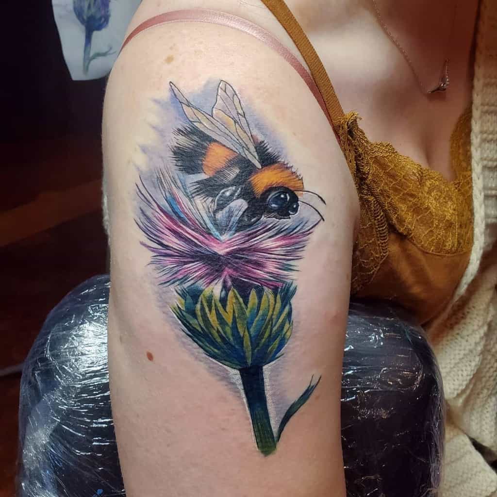 Vibrant tattoo of a bumblebee perched on a colorful thistle flower, located on the upper arm