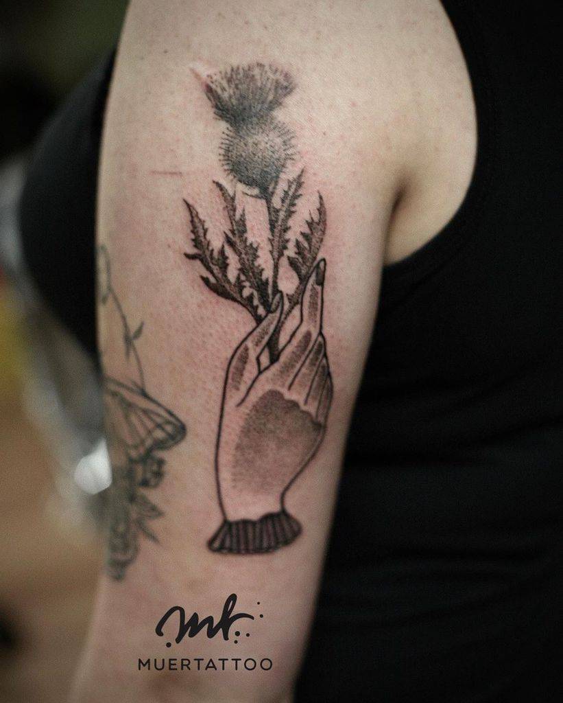 A beautifully intricate tattoo of a hand gracefully cradling a vivid thistle flower adorns the person's arm, capturing the essence of nature's resilience and adding a touch of wild elegance to their skin