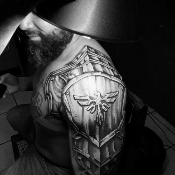 Realistic black and grey upper arm tattoo of a Hylian Shield from The Legend of Zelda, integrated into armor plating for a full shoulder piece look.