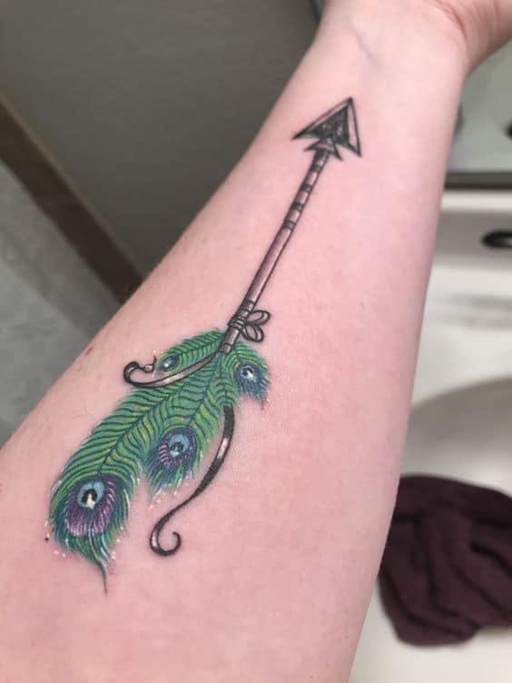 Peacock feather and arrow tattoo on the forearm, featuring vibrant green and blue hues with fine details and intricate shading.