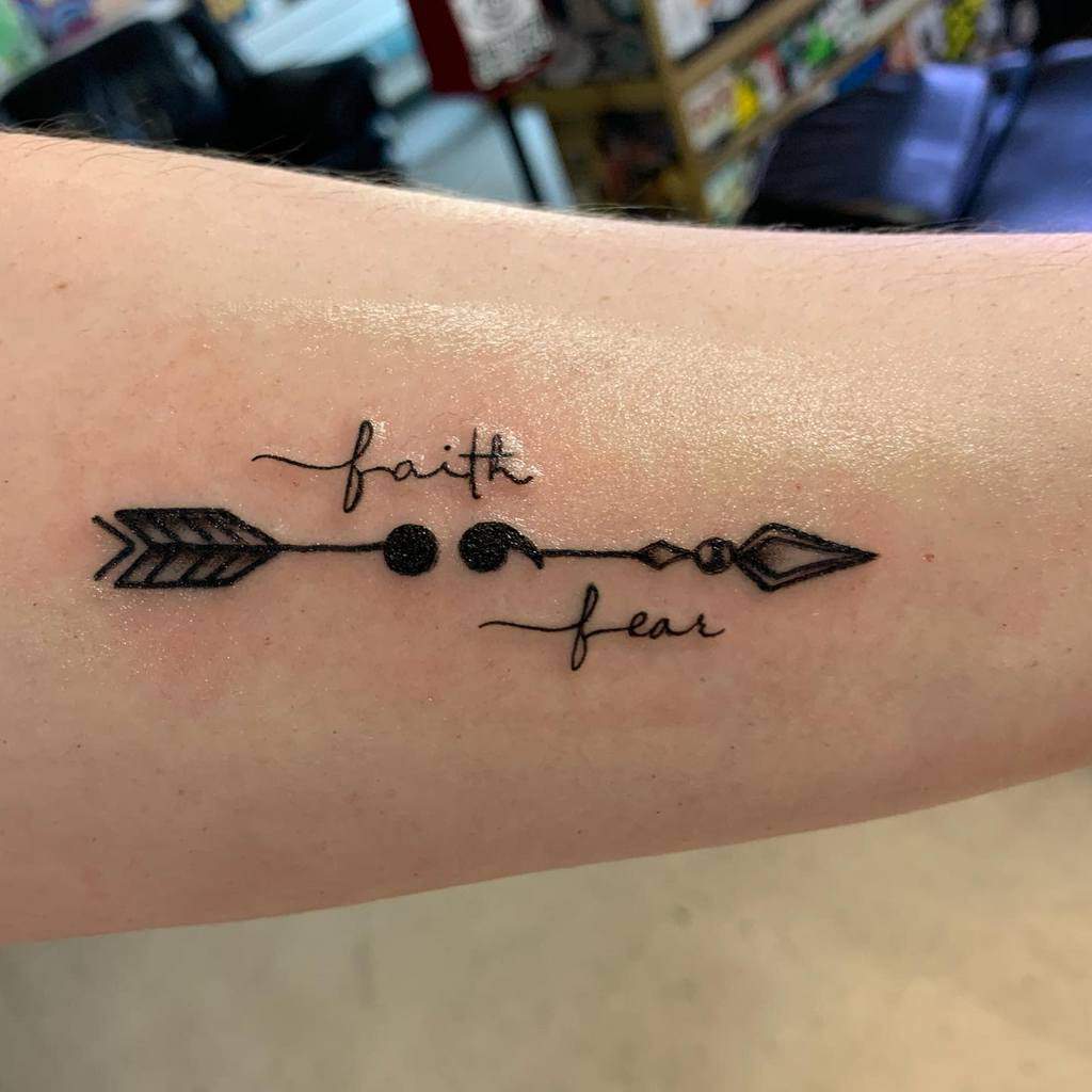 The tattoo features the words "faith" and "fear" linked by an arrow, with decorative elements creating a symbolic representation of balance and direction.