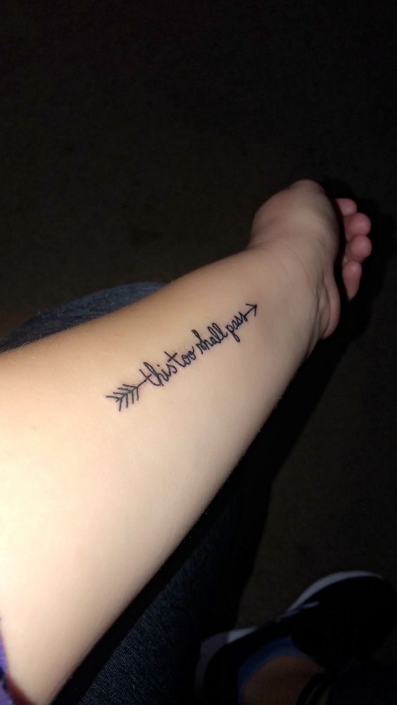 A "This Too Shall Pass" tattoo in cursive with arrows at both ends, on a person's forearm.