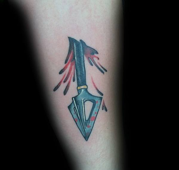 Bold broadhead arrow tattoo with red blood splatter effect, giving a dramatic and edgy look on the forearm.