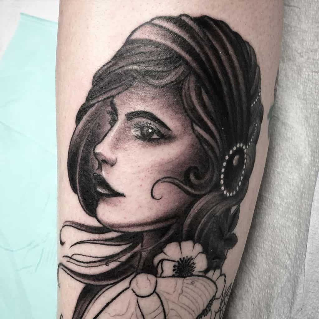 Black and grey tattoo of a gypsy woman with intense eyes, flowing hair, and floral accents, featuring bold shading on the leg.