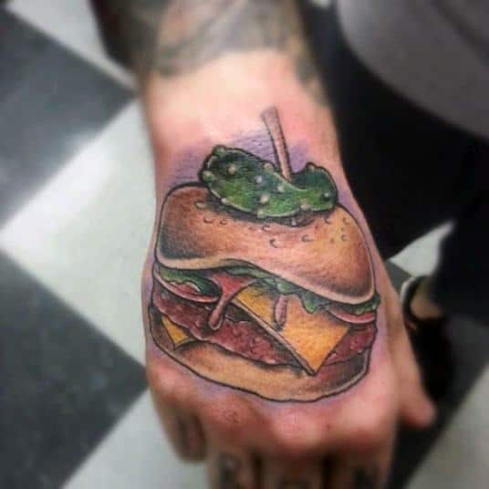 Cheeseburger tattoo with a slice of pickle, inked on a person's hand