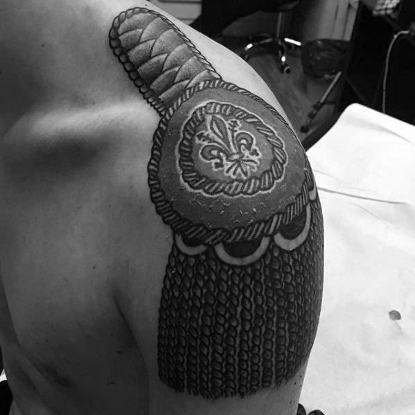 Detailed epaulette tattoo on shoulder with braided textures, scalloped edges, and a central fleur-de-lis emblem.