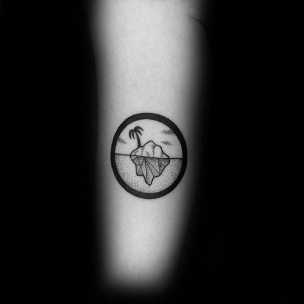 Small circular iceberg tattoo on the forearm, with a palm tree and ocean scene above and detailed underwater portion below.